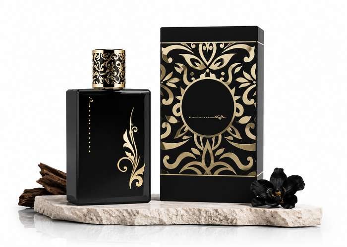 Black perfume bottle and matching fragrance box with elegant gold floral patterns, displayed on a marble slab with wood and black flowers.